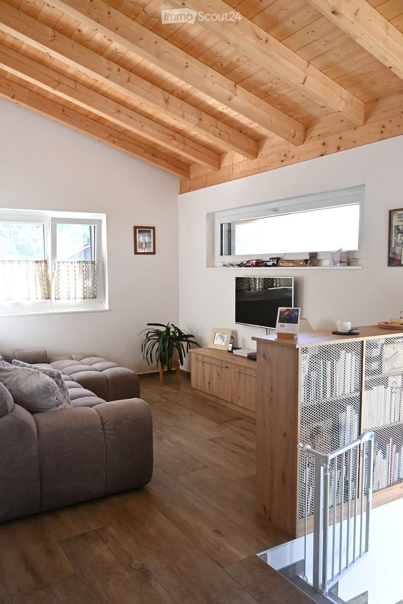 Bright living room with wooden ceiling, large windows, grey sectional sofa, coffee table, plant, TV, and decorative items on shelves.