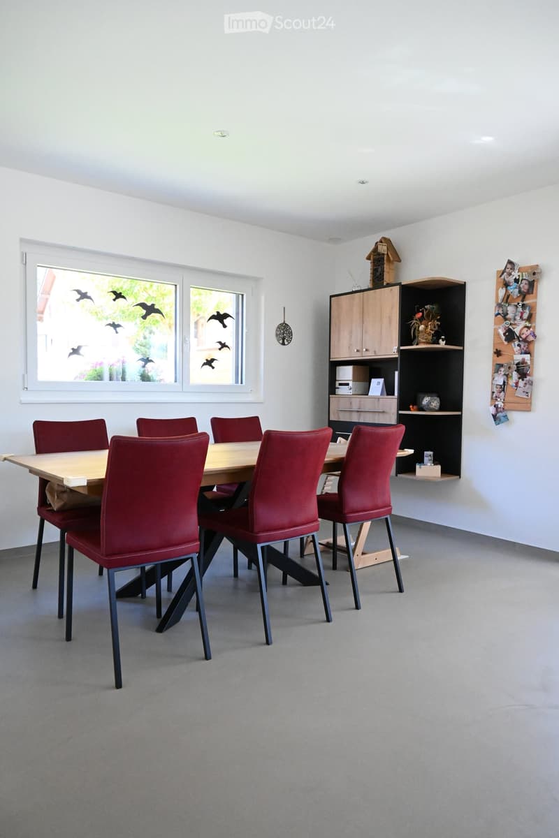 Bright dining room with a wooden dining table surrounded by red chairs, a cabinet, and a shelf with decorative items.