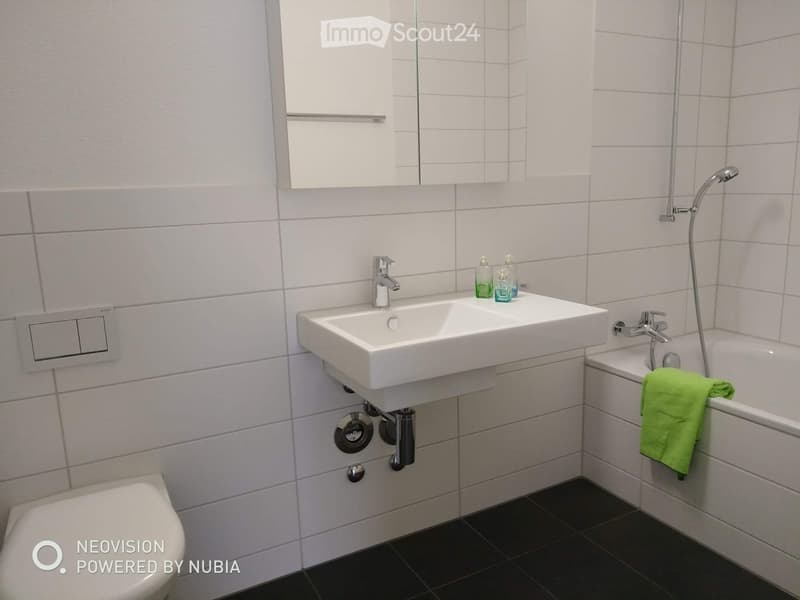 Modern bathroom with white tiles, a double sink, a bathtub, a toilet, a mirror, and a green towel on the side of the tub.