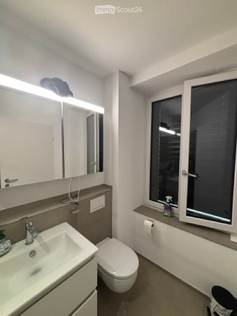 Modern bathroom with a large mirror, sink, toilet, and window. Features a sleek design with white cabinets and a light fixture above the mirror.