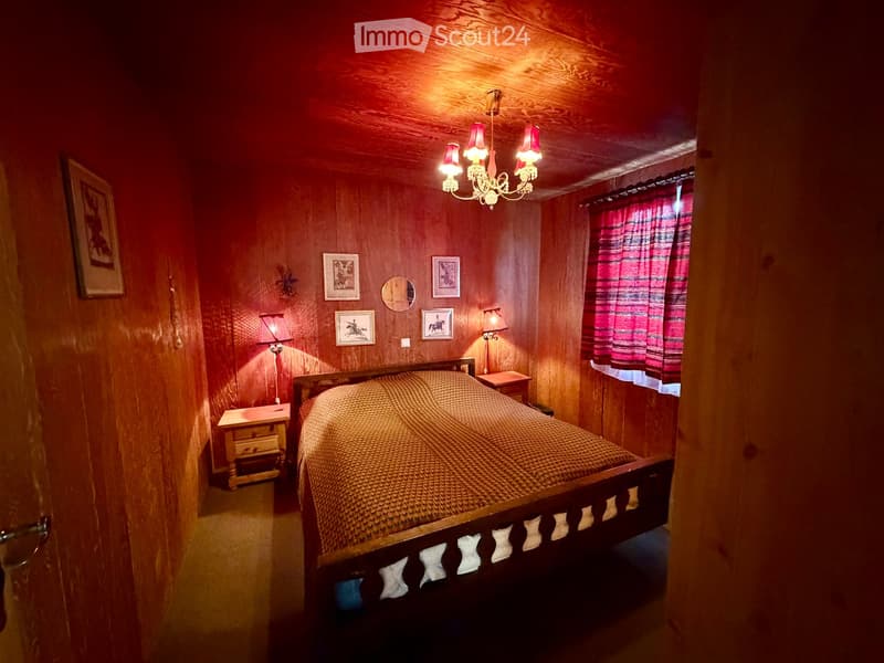 Cozy bedroom with wooden walls and ceiling, featuring a large bed with a woven bedspread, nightstands with lamps, framed pictures on the walls, and a chandelier with red shades.