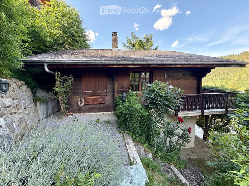 A rustic wooden house with a stone foundation, surrounded by lush greenery and a well-maintained garden. The house features a wooden balcony and a chimney, with a stone wall and a pathway leading to the entrance.