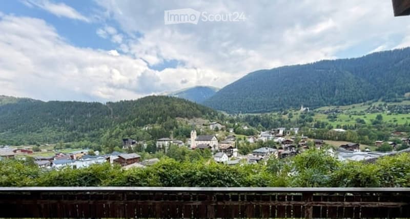 Panoramic view of a picturesque village nestled in a lush green valley surrounded by mountains, with scattered houses and a church visible.
