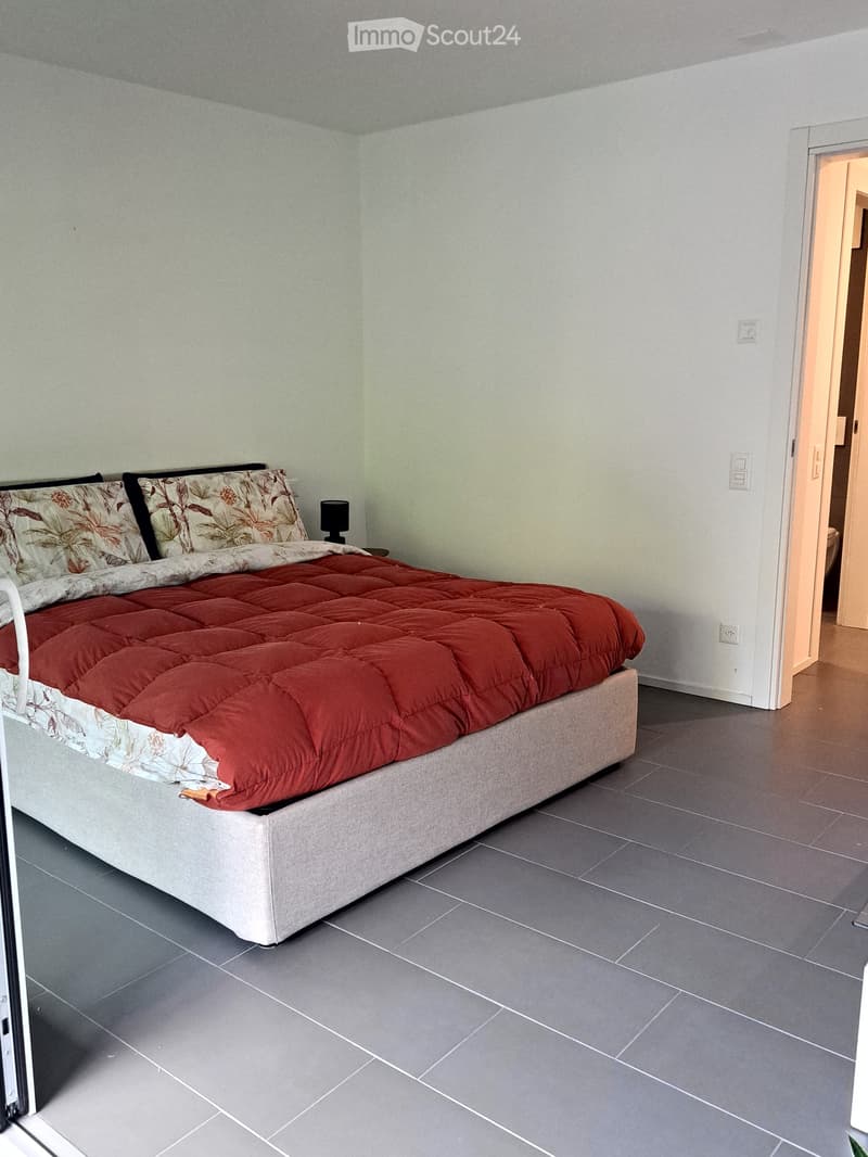 Modern bedroom with a large bed, red comforter, floral pillows, nightstand, and lamp. Grey tiled floor and white walls.