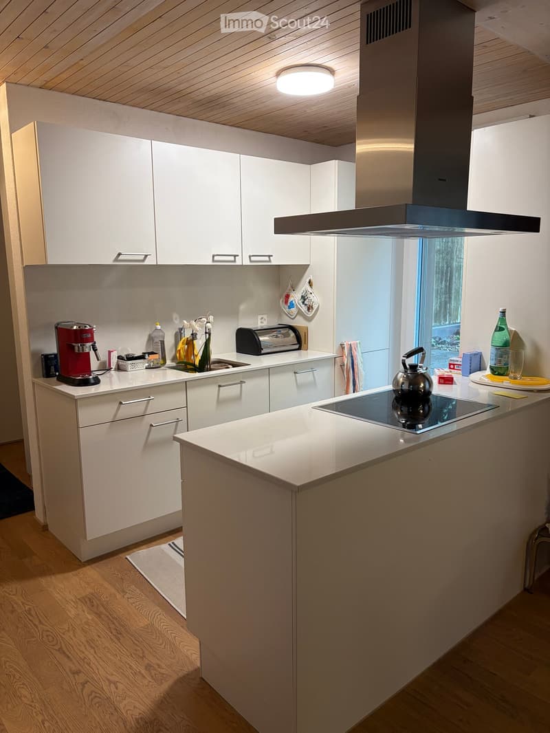 Modern kitchen with white cabinets, wooden flooring, and a central island with an induction stove. Features include a coffee maker, toaster, teapot, and various kitchen items on the countertops.