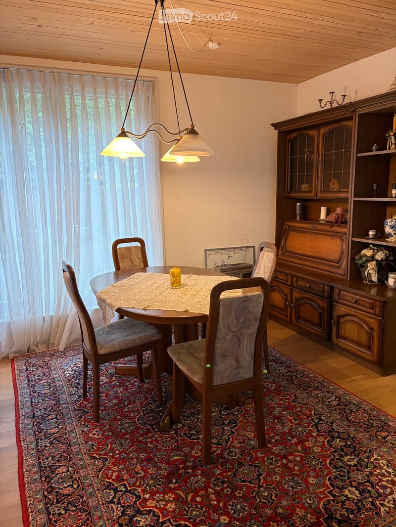 Dining room with a round wooden table, four chairs, a large wooden cabinet with glass doors, a flower vase, and a candle on the table. The room features a patterned rug, wooden flooring, and large windows with white curtains.