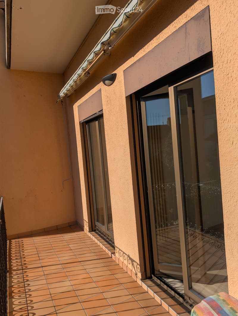 Balcony with tiled floor, glass sliding doors, beige walls, and a small awning above.
