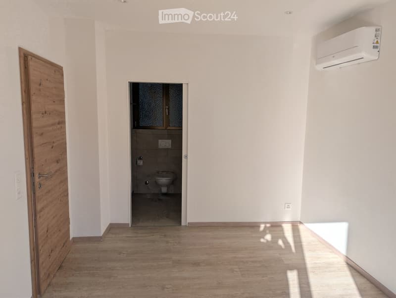 Empty room with light wooden flooring, white walls, and a wooden door leading to a small bathroom with a toilet and sink.