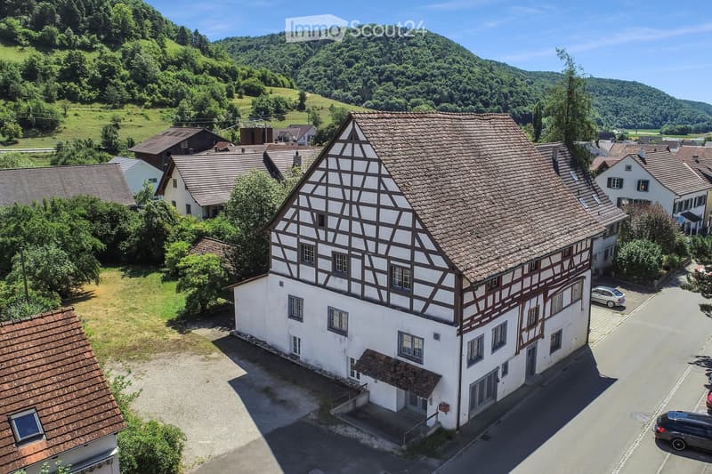 A traditional half-timbered house with a tiled roof, surrounded by other houses and greenery in a rural setting.