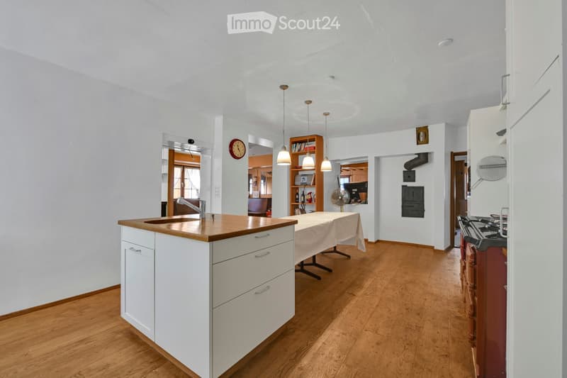 Bright kitchen with white cabinets, wooden countertop, and hardwood flooring. Features a sink, faucet, table, fan, oven, bookshelf, and clock.