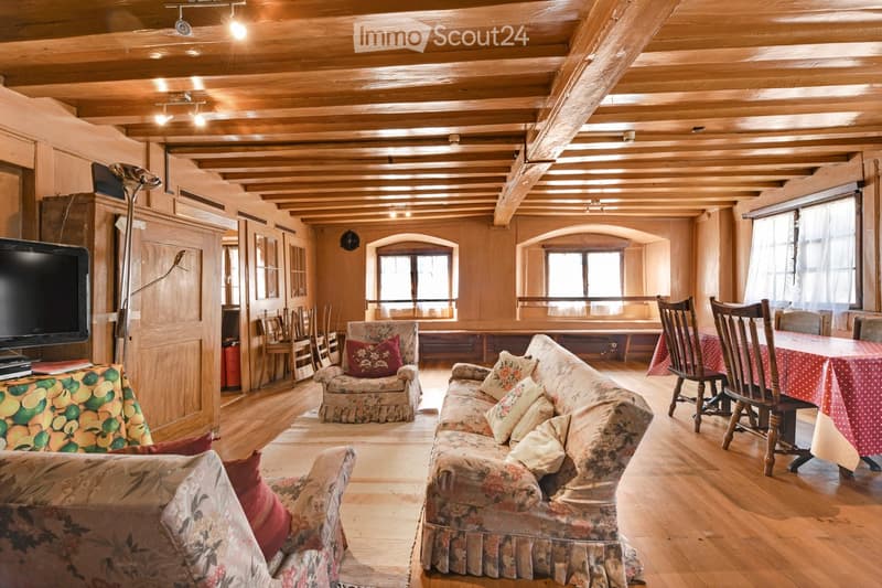 Cozy living room with wooden beams, exposed wooden ceiling, large windows, floral-patterned sofa and armchair, dining table with chairs, television, and floor lamp.