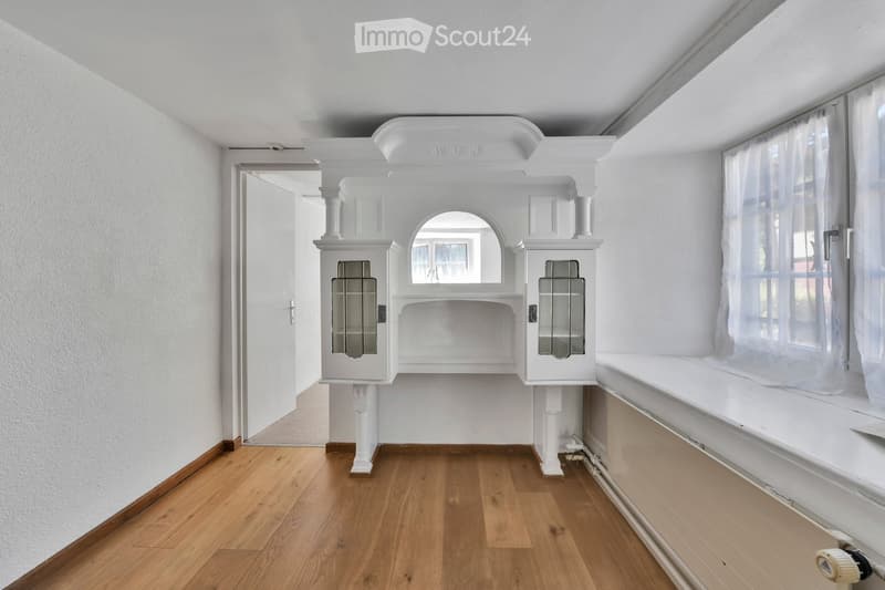 Empty room with wooden flooring, white walls, built-in white cabinetry with glass doors, and a large window with white curtains.