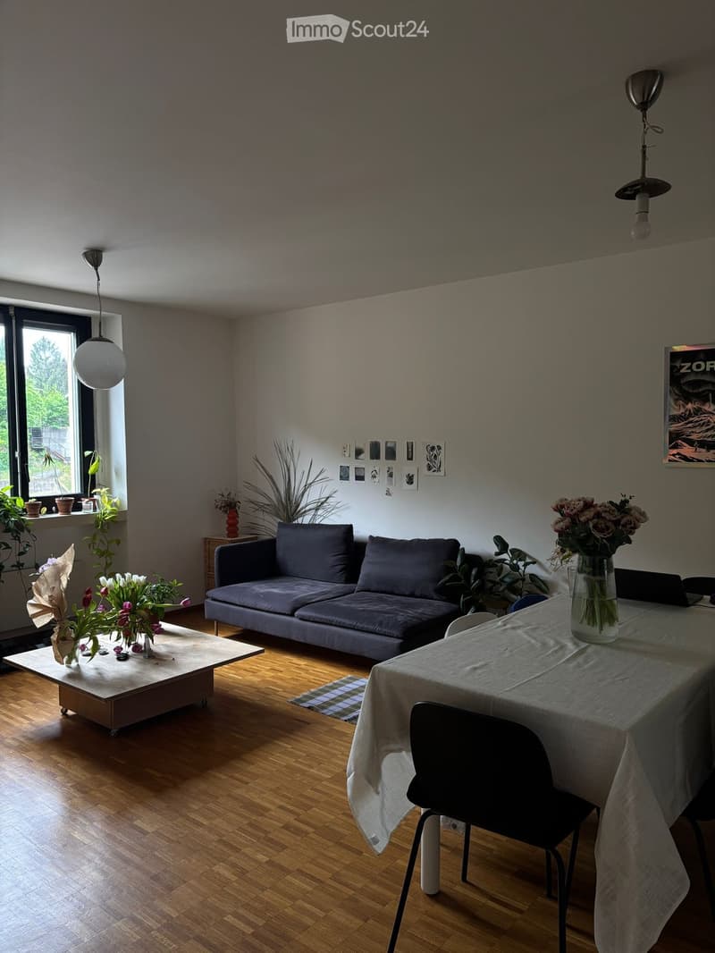 Modern living room with wooden flooring, grey sofa, coffee table, dining table with chairs, and decorative vases with flowers.