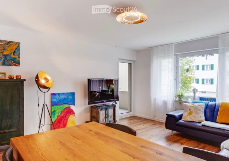 Bright living room with wooden flooring, large windows, a blue leather sofa, a wooden dining table, a television, and decorative artwork.