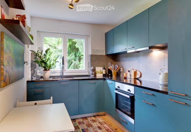 Modern kitchen with blue cabinets, stainless steel appliances, a sink, and a small dining area with a table and chair. A potted plant and a painting add a touch of decor.