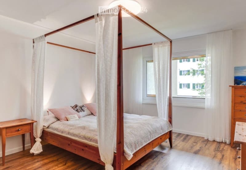 Cozy bedroom with a wooden four-poster bed, nightstand, dresser, and a painting on the wall. The room features wooden flooring and large windows with white curtains.