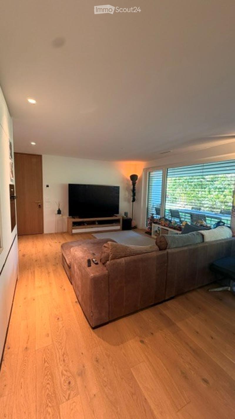 Modern living room with light wooden flooring, large windows offering a view, a grey sectional sofa, a coffee table, a television on a stand, and a floor lamp.