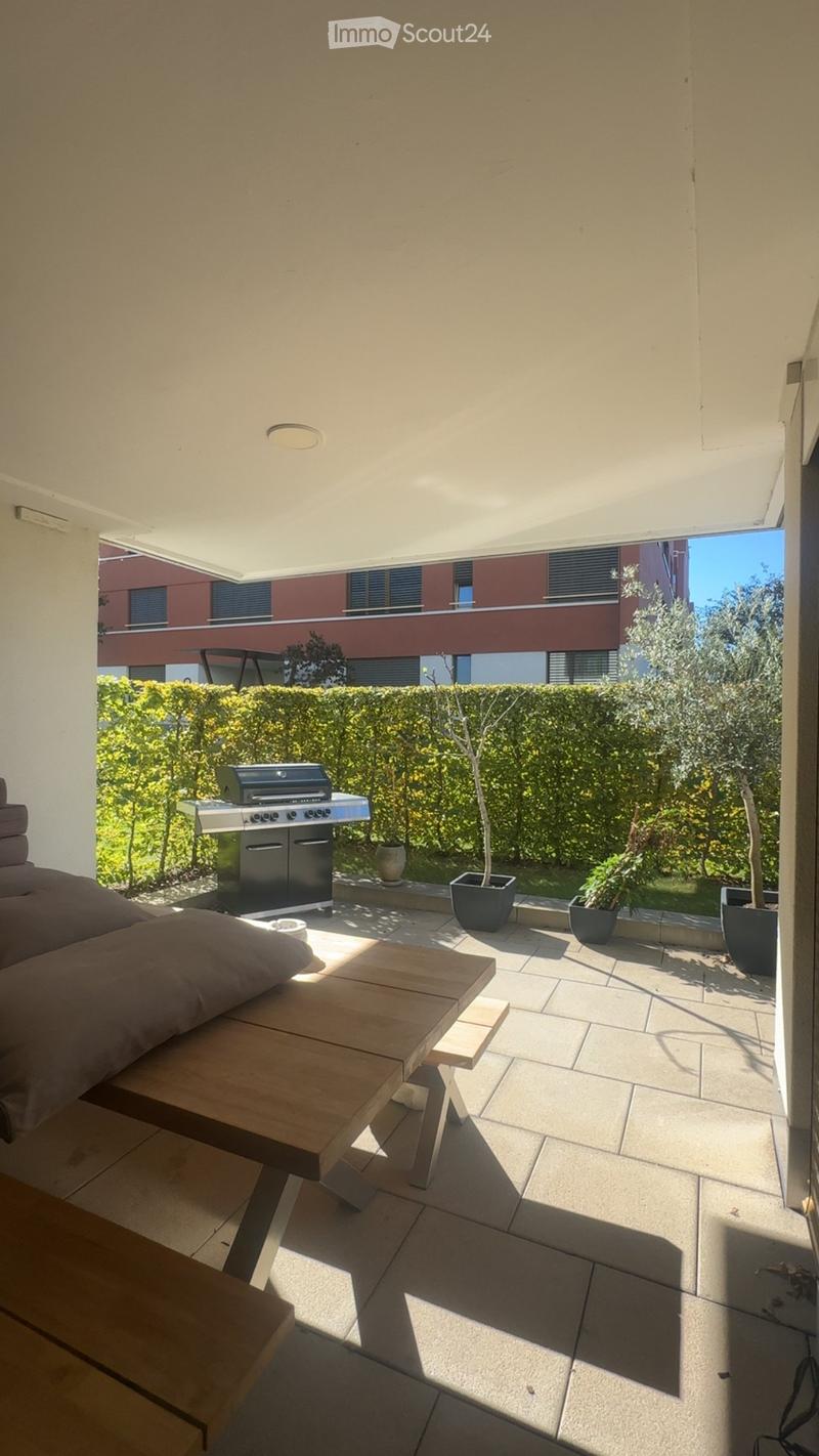 Balcony with a wooden table, sofa, grill, and several potted plants, overlooking a landscaped garden area.