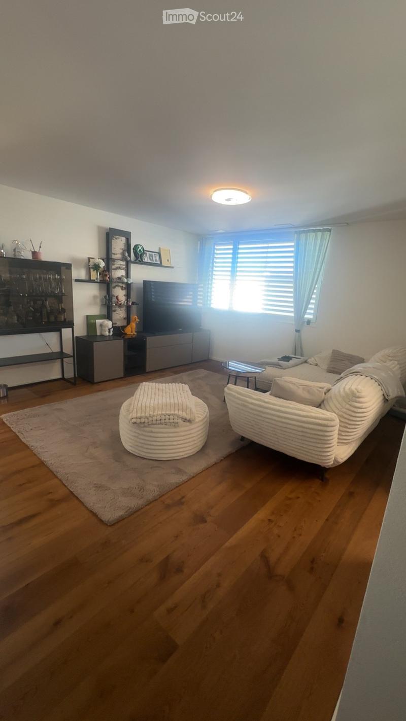 Modern living room with wooden flooring, large window with curtains, sofa, coffee table, ottoman, TV, cabinet, and shelves.