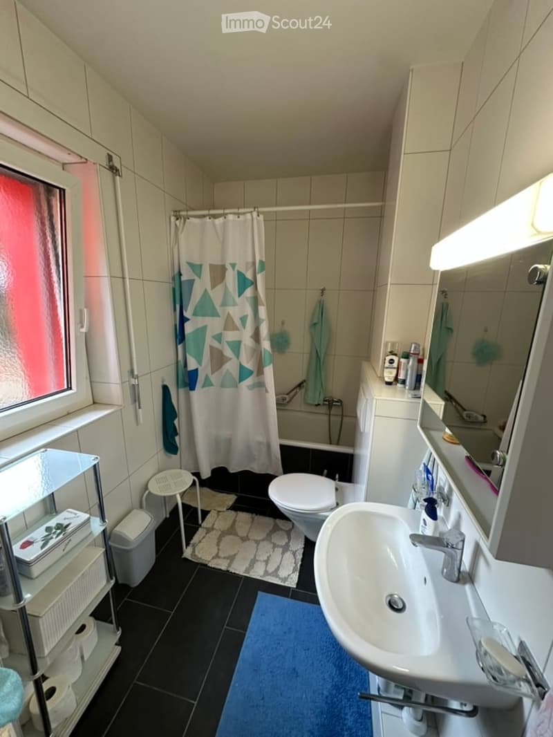 Modern bathroom with white tiles, a large mirror, a sink, toilet, shower, and a small stool. Features a colorful shower curtain and a towel rack with towels.