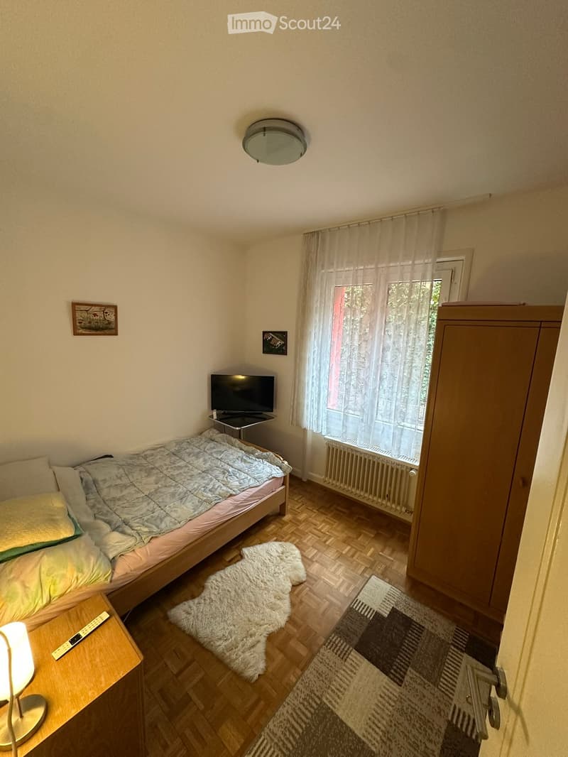 Cozy bedroom with wooden flooring, a single bed with a grey duvet, a nightstand with a lamp, a television, a fluffy white rug, and a small picture frame on the wall.