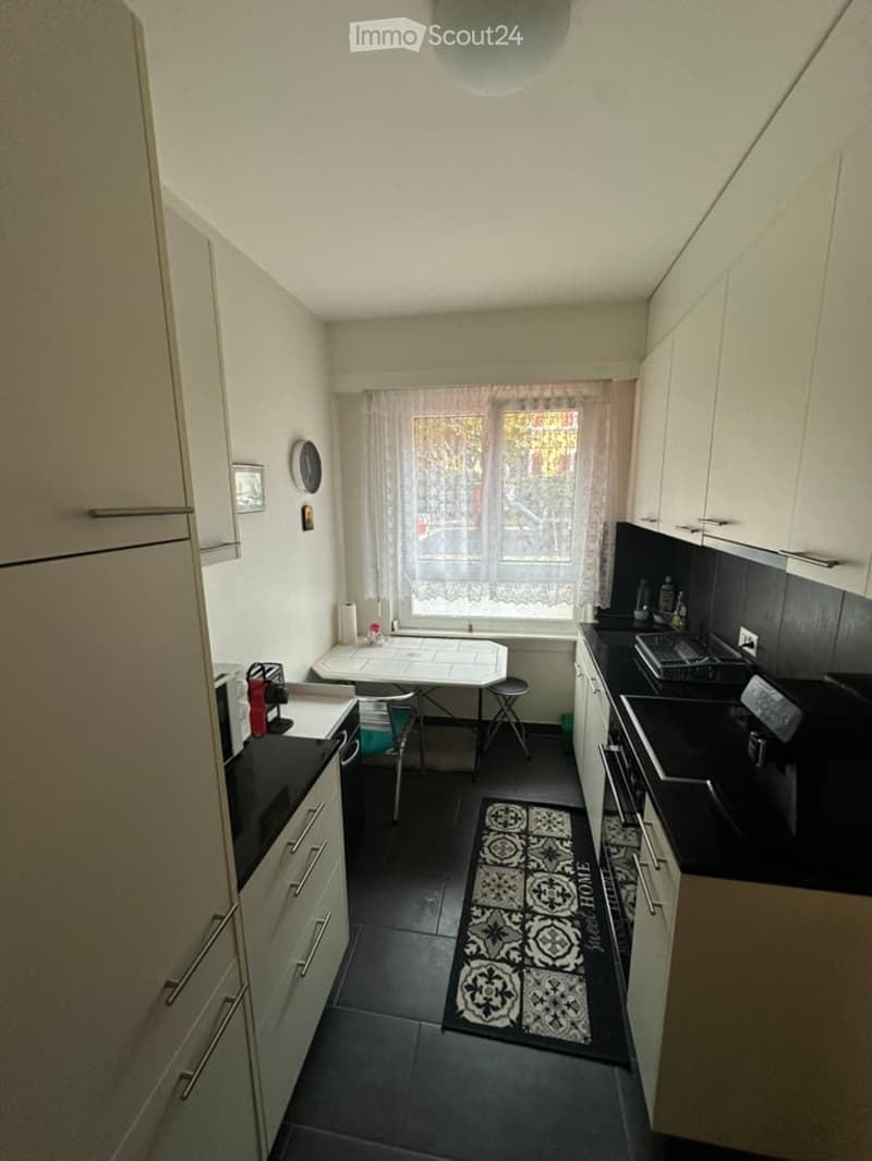 Modern kitchen with white cabinets, black countertops, and a small dining area. Features include a window with sheer curtains, a black and white patterned rug, and various appliances.