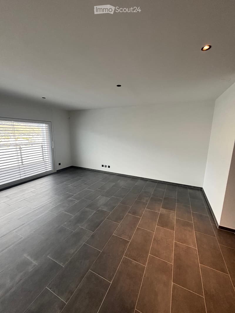Empty room with grey and brown tiled floor, white walls, large window with blinds, and recessed lighting.