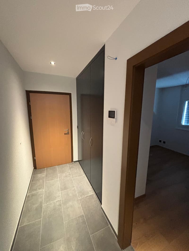 A narrow hallway with tiled flooring, white walls, and two doors: one wooden and one glass-paned. A small light fixture is mounted on the ceiling.