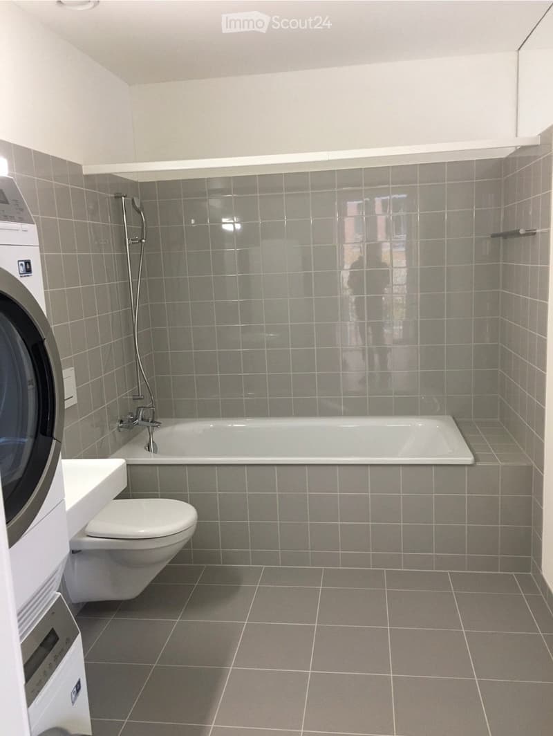 Modern bathroom with grey tiled walls and floor, featuring a bathtub, toilet, and washing machine.