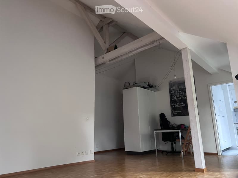 A modern, open-concept room with exposed wooden beams, a refrigerator, a desk with a chair, and a clock on the wall.