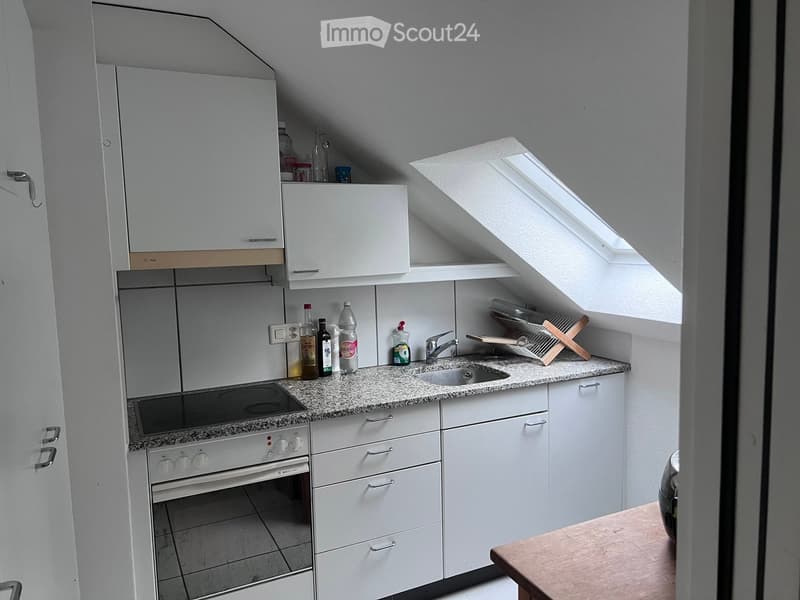 Compact kitchen with white cabinets, granite countertop, sink, oven, and various bottles on the counter.