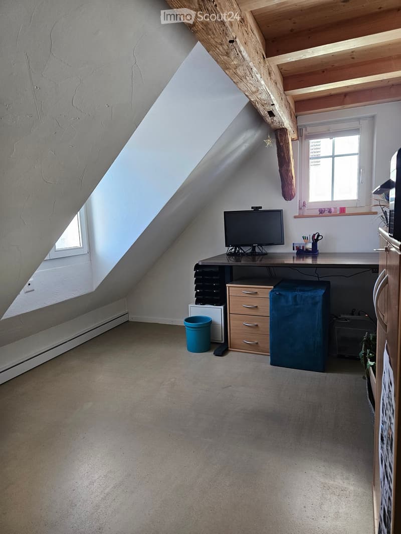 Attic room with slanted wooden ceiling, large windows, desk with screen, chair, and cabinet.