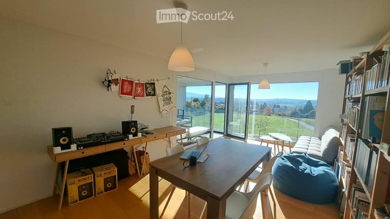 Bright living room with large windows offering a scenic view, featuring a sofa, beanie chair, dining table, chairs, laptop, bookshelf, speakers, and artworks.
