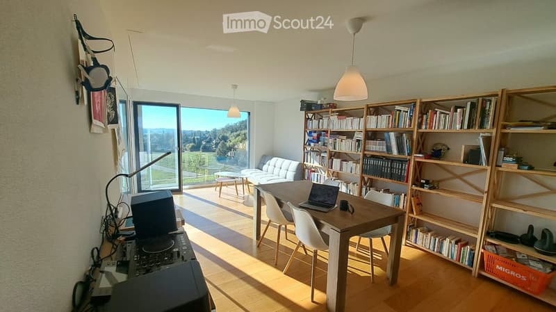 Bright, open living space with wooden flooring, large windows offering a scenic view, a sofa, a dining table with chairs, a laptop, and a bookshelf filled with books.