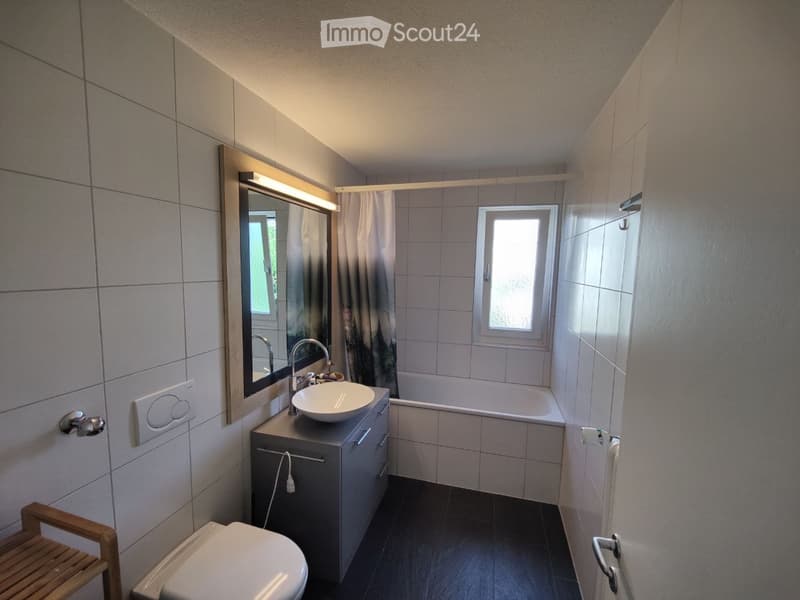 Modern bathroom with a white sink, bathtub, toilet, and large mirror. The room features tiled walls and a window providing natural light.