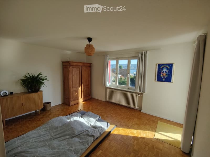 Bright bedroom with wooden flooring, large window with view, bed, wardrobe, cabinet, plant, picture frame, and wicker basket.