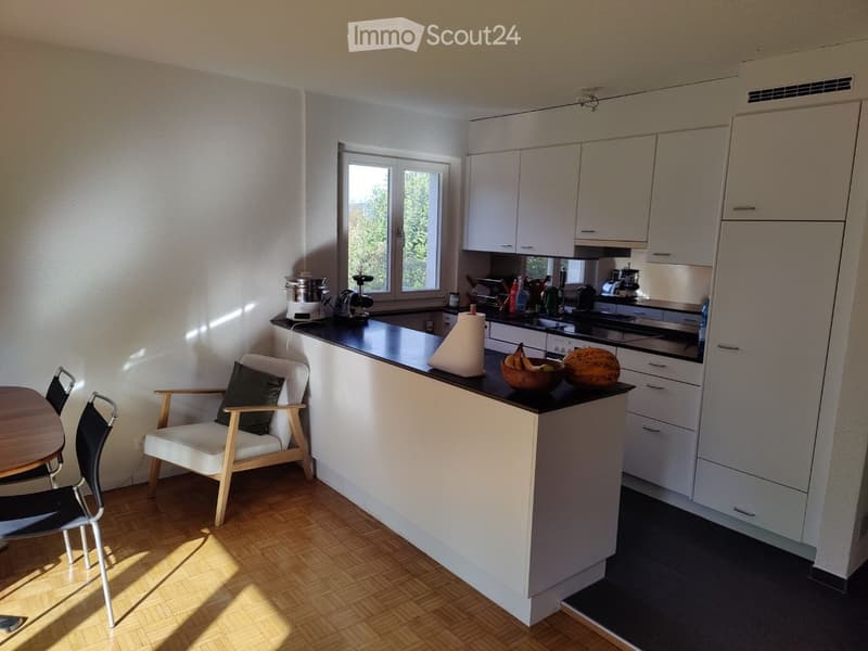 Modern kitchen with white cabinets, black countertops, and a small dining area featuring a chair and table. The kitchen includes various appliances and a bowl of fruit on the counter.