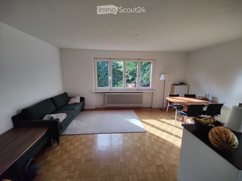 A well-lit living and dining area with wooden flooring, a dark green sofa, a dining table with chairs, and a floor lamp. The room features large windows with a view of greenery outside.