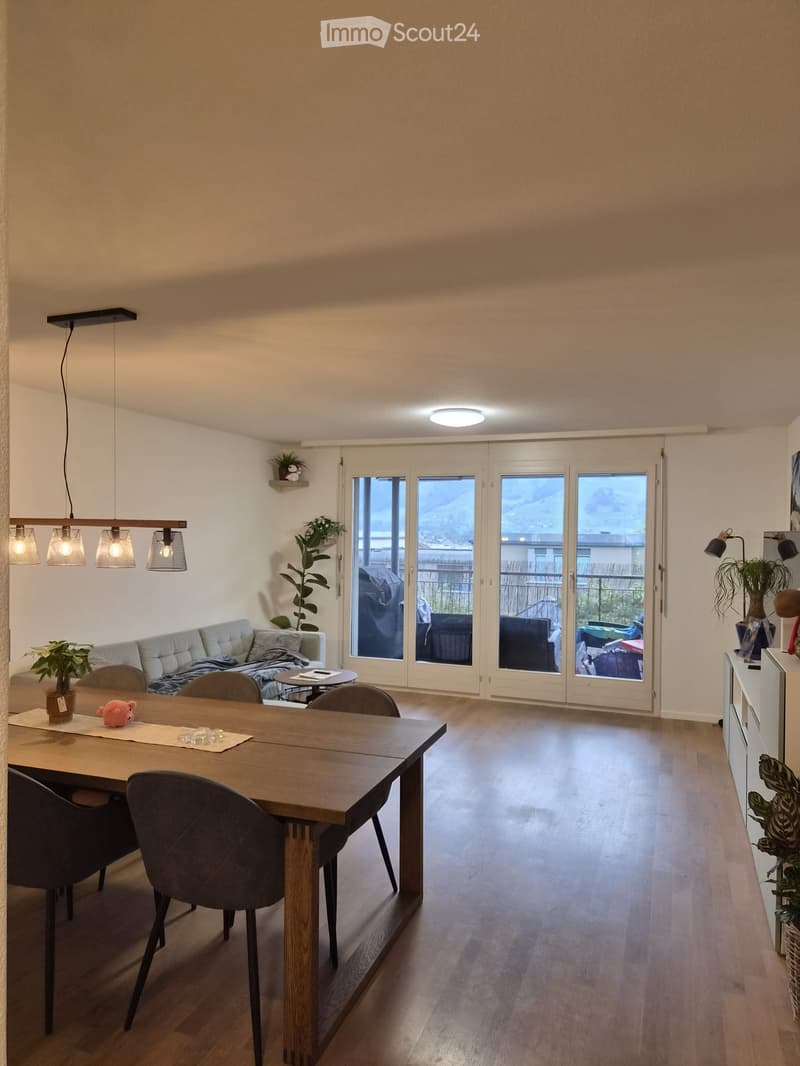 Bright living and dining area with wooden flooring, large sliding glass doors leading to a balcony, a sofa, dining table with chairs, pendant lights, and plants.