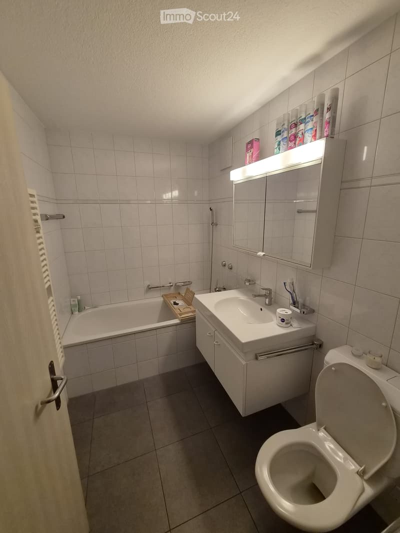 Modern bathroom with white tiles, a bathtub, sink, toilet, mirror, and a built-in cabinet with a light fixture above the mirror. The sink has a faucet and toothbrush holder, and the bathtub has a wooden tray with items on it.