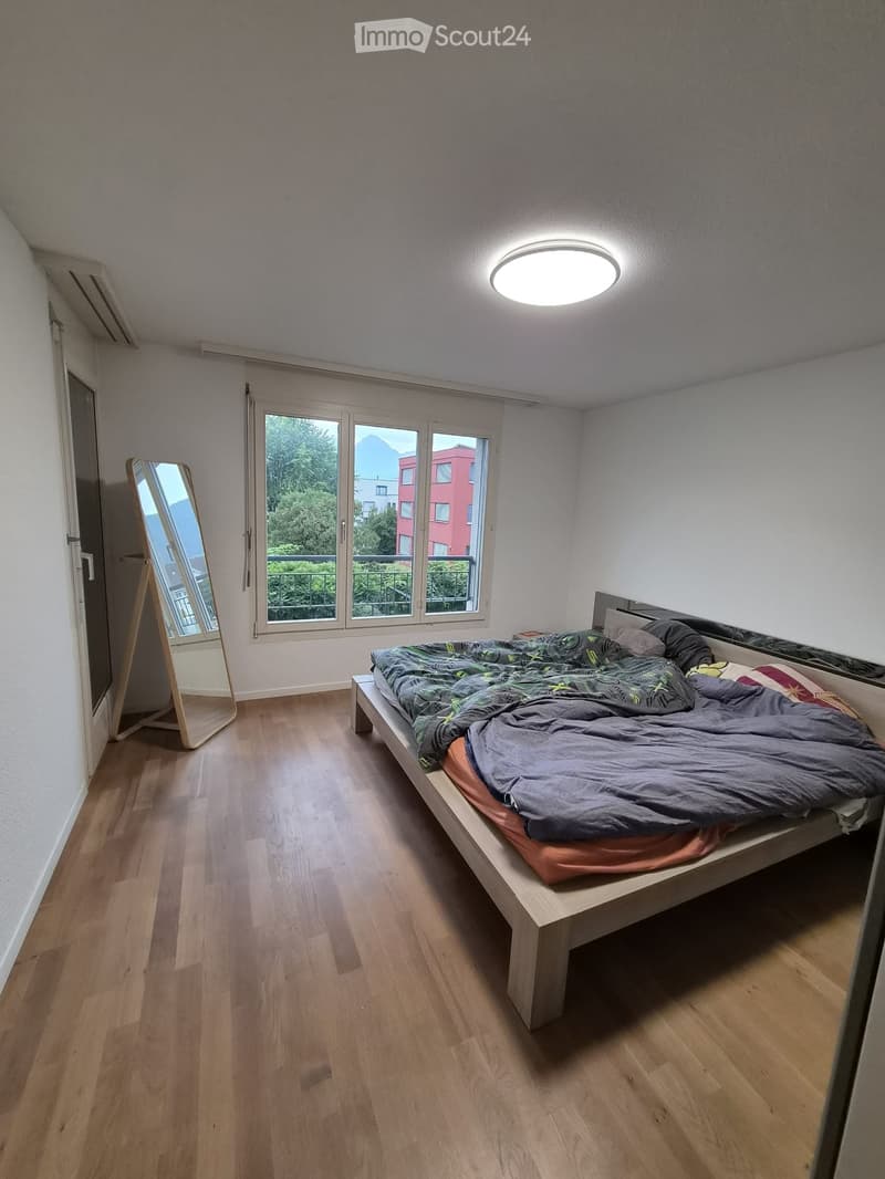 Bedroom with wooden flooring, a single bed with a grey duvet, a standing mirror, and large windows offering a view of trees and buildings.