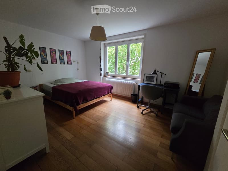 Cozy bedroom with wooden flooring, a bed with a purple duvet, a desk with a chair, a sofa, a large mirror, and a potted plant near the window.