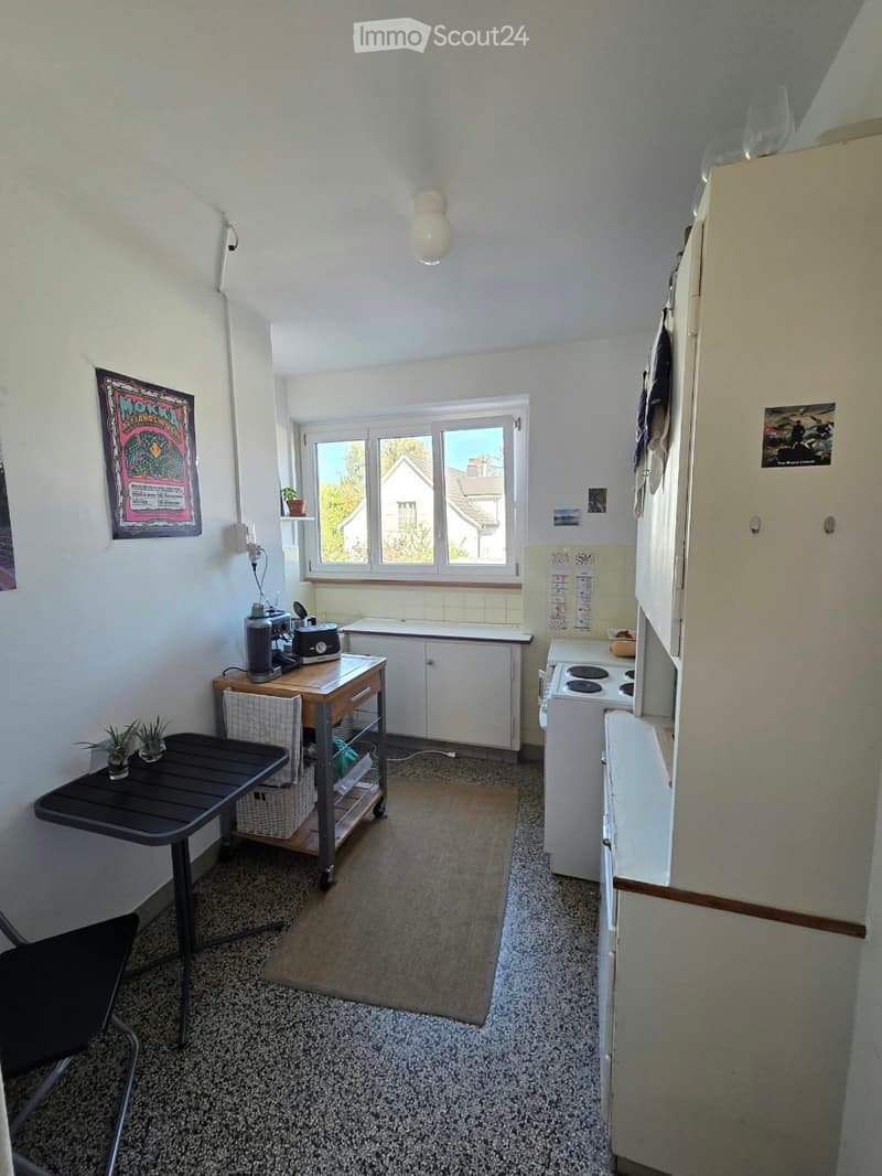 A compact kitchen with white cabinets, a refrigerator, a stove, a small table with chairs, and a coffee maker on a wooden counter. There is a window with a view of the outside, and a potted plant on the table.