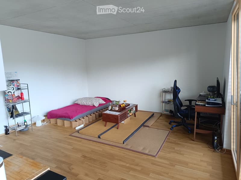 A small, furnished room with a tatami mat area, a bed with pillows, a desk with a chair, and a shelf with various items.