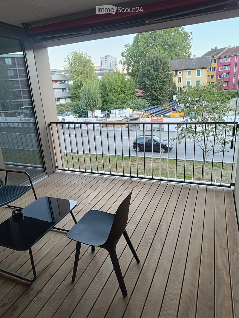 Balcony with wooden decking, black chairs, and a small table, overlooking a street with passing cars and buildings.