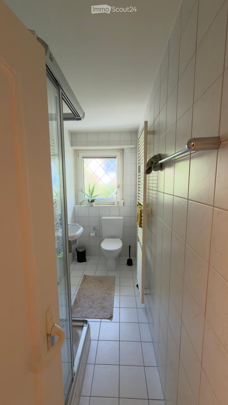 A small, well-lit bathroom with white tiled walls and floor, featuring a toilet, sink, bathtub, towel rack, and a small plant on the windowsill.