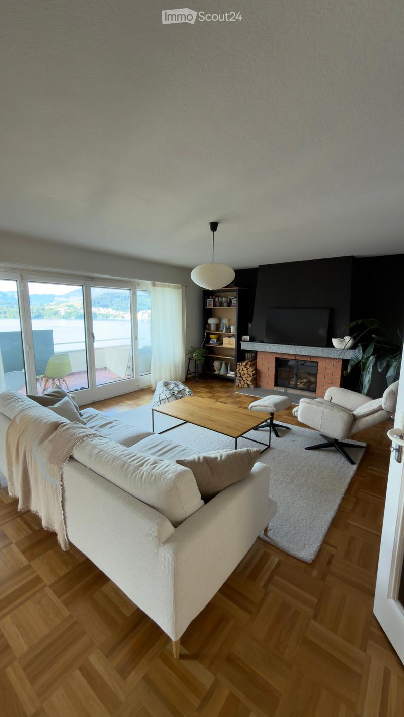 Modern living room with wooden flooring, large windows offering a scenic view, a beige sofa, coffee table, fireplace, bookshelf, and a television.