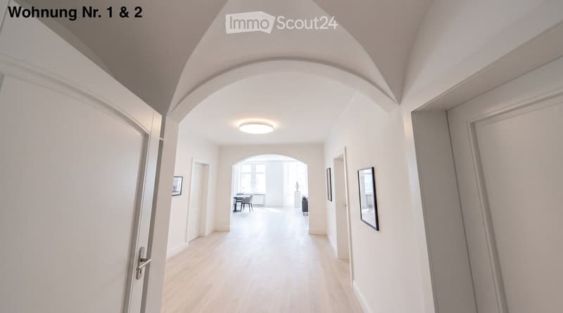 Bright hallway with wooden flooring, white walls, and recessed lighting. Features arched doorways leading to adjacent rooms, with visible furniture in the background.