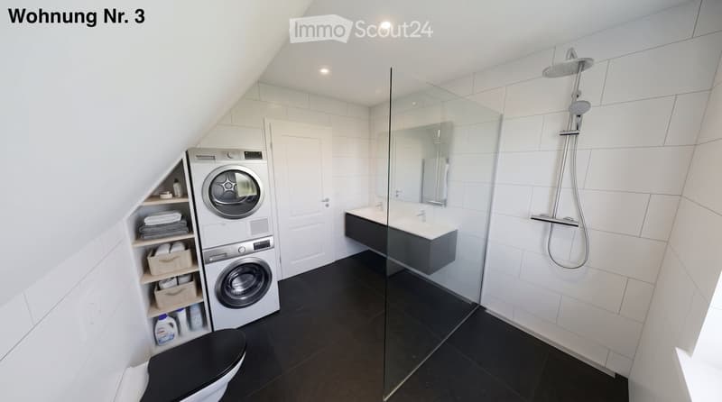 Modern bathroom with a toilet, shower, sink, mirror, and a private washing machine. The space features white tiled walls, a black tiled floor, and a glass-enclosed shower area.