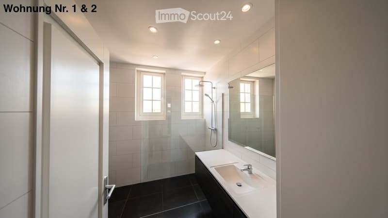 Modern bathroom with white tiles, black floor, large mirror, sink, and glass-enclosed shower area.